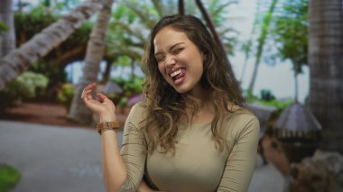Ağaç ve tropikal çevresiyle yemyeşil bir parkta gülümseyen genç İspanyol kadın neşeli ifadeler ve günlük giysiler sergilerken güneşli havanın tadını çıkarıyor..
