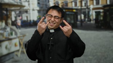 Şehir ortamında gülümseyen rahip gözlük takıyor ve rahip tasması takıyor. İki eliyle bir kaya sembolü yapıyor..