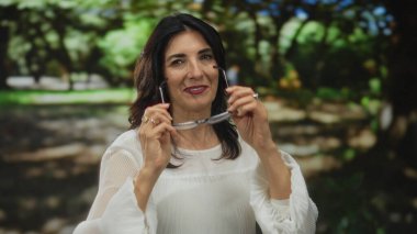 Beyaz bluz giyen orta yaşlı bir kadın yeşil park ortamında gözlük ve parmaklarını düzeltiyor; kendine güven huzuru.