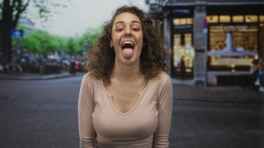 Çarmıha gerilmiş kolları ve dili ile gülümseyen genç İspanyol kadın şehir caddesinde dükkan önlerinde ve park edilmiş bisikletlerin yanında;.