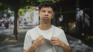 Gözlüklü adam, şehir sokağında ağaçlarla kaplı beyaz gömleğini çekiştiriyor ve yaz güneşinin altında park edilmiş arabaları çekiyor; rahatsızlık verici..