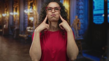 Kırmızı gömlekli ve kırmızı gözlüklü bir kadın binada şişmiş yanakları olan bir heykeli işaret ediyor..