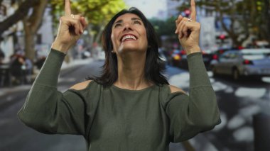 Orta yaşlı İspanyol kadın, güneşli şehir caddesinde parmaklarını yukarı kaldırarak gülümsüyor; umut iyimserliği.