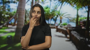 Kadın parmakları ve katlanmış kolları ile çene dokunuyor, genç bir ormanda siyah gömlek giyiyor palmiyeler ve yeşil çimlerle kaplı; düşünceli bir yansıma.