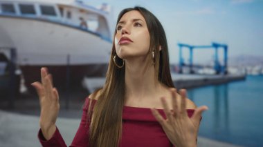 Güneşli bir sahil limanında rıhtımdaki teknelerin yanında bordo bir bluz giyerken uzun saç fırçalayan kadın; özgüven zarafeti.