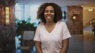 Kadın otelin lobisinde geniş bir gülümseme içinde oturuyor ve bitkiler, eller rahat ve kameraya bakan vücut; sevinç bağlantısı sıcaklığı.