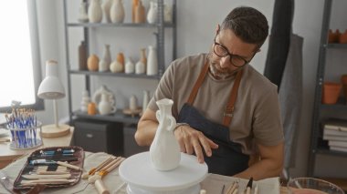 Önlüklü bir adam stüdyoda çömlekçi çarkında elle beyaz seramik vazoyu şekillendirir fırçalar, aletler ve tencere raflarıyla çevrelenir; sanatsal odaklanma.