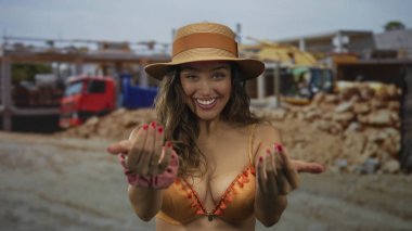 Güneş ışığı altındaki inşaat binasında elleri turuncu bikinili bir kadın çağırıyor; eğlenceli bir davet.