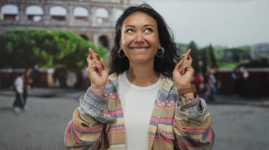 Parmakları çapraz olan ve Roma kolezyum binasına bakan, gülümseyen, renkli desenli ceket giyen ve halka küpeler takan bir kadın; umut yolculuğu dileği.