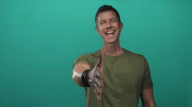 El sıkışmak için sağ elini uzatan ve zeytin tişörtü ve bilezikleriyle bir deniz mavisi stüdyosunda gülümseyen adam; dostça karşılama.