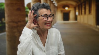 Gri saçlı yaşlı bir kadın, eli kulağında bir jest yapıyor, eski bir kasaba caddesinde gülümsüyor, şehir çevresi ile iç içe, açık bir atmosferi vurguluyor..