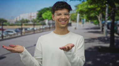 Yuvarlak gözlüklü ve beyaz bluzlu bir adam gülerken ağaçlık şehir sokağında avuçlarını açık tutar; belirsizlik..
