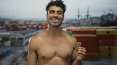 Genç İspanyol adam çıplak göğsü olan bir granola bar tutuyor ve renkli nakliye konteynırı binasında kameraya gülümsüyor. Mutluluk..