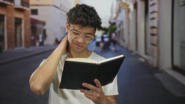 Şehrin güneşli caddesinde eli çenesinde kitap okuyan, gözlüklü ve beyaz tişörtlü bir adam; merak yansıması.