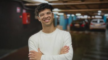 Kapalı otoparkta arabasının yanında kollarını kavuşturmuş gözlüklü ve kazaklı genç bir adam. Kendine güveniyor..