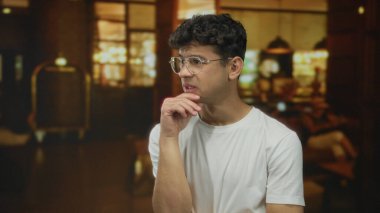 Beyaz gömlek giyen ve gözlüklü bir adam lobide sıcak ışıklarla çeneye dokunuyor..