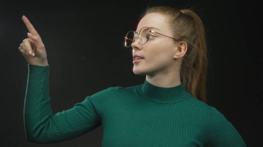 Stüdyoda parmağını kaldırmış, işaret parmağını kaldırmış, yana bakarken, gözlük ve yeşil süveter takan genç bir kadın..
