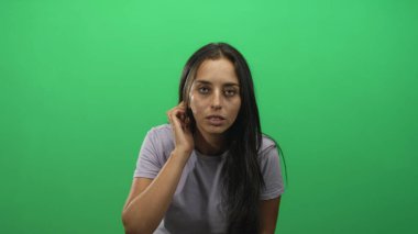Genç İspanyol kadın, yeşil stüdyoda oturmuş kameraya bakarken eliyle kulağına dokunuyor..