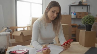 Yeni evde telefon kullanırken notlar yazan bir kadın. Etrafı kutular ve ahşap masada saksı bitkileriyle çevrili..