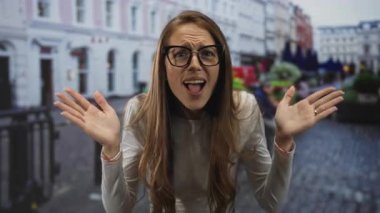 Elleri havada, şehir sokaklarında, gözlüklü, beyaz gömlekli, ağzı açık ve kaşlarını kaldırmış bir kadın..