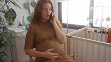 Hamile kadın telefonu kulağına tutuyor ve bir binanın bebek odasında rahatça otururken çıplak karnını kucaklıyor; annelik beklentisi.