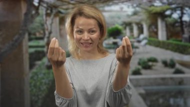 Genç sarışın kadın para için parmaklarını çimdikliyor güneş ışığı altında binanın önünde hareket ediyor; beklenti.