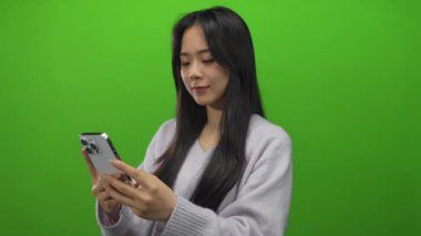 Genç Çinli kadın izole edilmiş yeşil arka plana karşı akıllı telefon kullanıyor modern teknoloji ve gündelik moda sergiliyor..