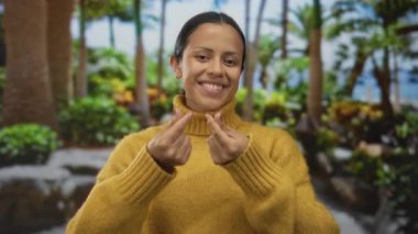 Hardal rengi kazaklı kadın parmaklarıyla kalp hareketi yapıyor yemyeşil bir parkta palmiye ağaçları ve açık hava ortamını güzelleştiren canlı bitkiler.