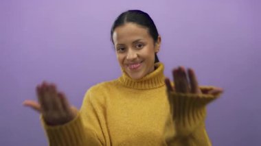 Genç İspanyol kadın canlı mor bir arkaplanda gülümsüyor. Sade sarı bir kazak giyiyor. Açık elleri var.