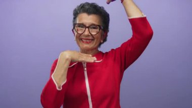 Kırmızı kazak giyen, gri saçlı ve gözlüklü son sınıf öğrencisi bir kadın, elleri mor bir arka plana dönük bir çerçeve hareketi yapıyor. Stüdyo ortamında neşe ve yaratıcılığını ifade ediyor..