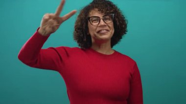 Woman with glasses wearing bright red sweater makes peace sign with right hand in studio portrait; joy.
