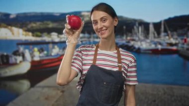 İskelede eldivenli kırmızı elmalı bir kadın önlük ve çizgili gömlek giyerken kameraya meyve sunuyor; sağlıklı beslenme sevinci.