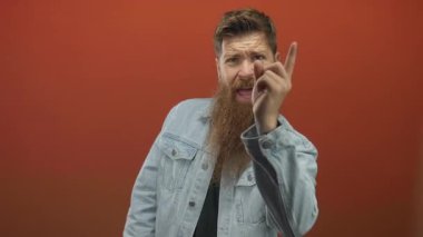 Sakallı adam kırmızı bir stüdyoda kamerayı işaret ediyor; meydan okumanın aciliyeti.
