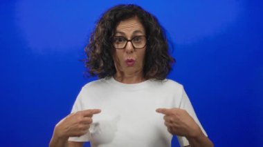 Orta yaşlı, gözlüklü İspanyol bir kadın izole edilmiş mavi bir stüdyo duvarının önünde parmaklarını göğsüne işaret ediyor..