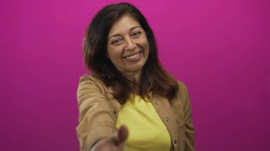 Sarı gömlekli kadın pembe stüdyoda kameraya doğru elini uzatıyor; güven bağlantısını selamlıyor.