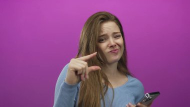 Akıllı telefon tutan genç beyaz kadın, mor stüdyoda küçük bir meblağ göstermek için parmaklarını çimdikliyor; kuşkulu ölçü boyutu minimalizm.