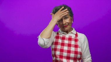 Önlüklü, gri saçlı yaşlı bir kadın mor arkaplan konusunda endişeli görünüyor. Kırmızı kareli önlük giyerken endişeli bir ifade takınıyor ve canlı bir ortamda duyguları ön plana çıkarıyor..