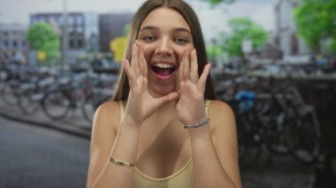 Güneş ışığı altındaki bir kanalda, kaldırım taşı sokağında elleri ile ağzını çerçeveleten bir kız arıyor; heyecan.