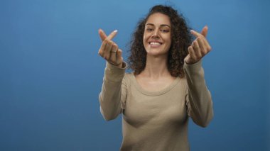 Mavi stüdyoda çıplak elle parmak kalp hareketi yapan kadın, kameraya neşeli bir ifadeyle gülümsüyor; neşe dostu sıcaklık.