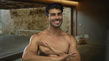 Genç İspanyol adam çıplak göğsünü gösteriyor, gülümsüyor ve el sallıyor. Sıcak ışık altında ahşap zemini olan boş stüdyo odasında, inkar ediyor..