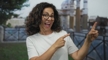 Orta yaşlı İspanyol kadın gülüyor ve Roma harabelerine işaret ediyor. Seyahat ve keşfin zevkini özetliyor..