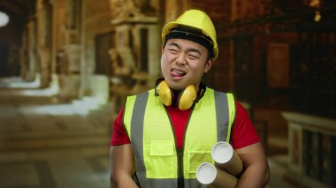 Korumalı yelek ve miğferli genç bir adam kilise koridorunun içinde, şakacı bir şekilde dilini uzatıyor..