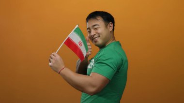 Elinde İran bayrağı tutan genç adam, başparmağıyla turuncu arka planda duruyor.