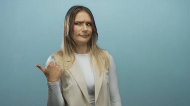 Sarışın kadın mavi bir arka planı işaret ediyor, şüphe ve merakını ifade ediyor, izole ve sıradan, beyaz ve bej giyiyor, düşünceli ifadesini vurguluyor..