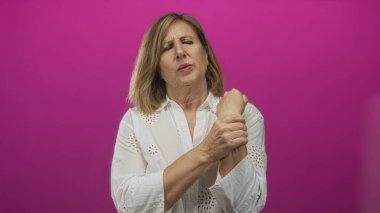 Beyaz bluzlu kadın bileğini kavrıyor ve stüdyoda pembe bir arkaplan önünde acı dolu bir ifadeyle ürküyor..