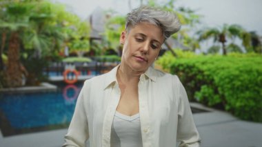 Beyaz gömlekli, gözleri kapalı ve başı eğik bir kadın binanın yanında mavi havuz kenarında duruyor..