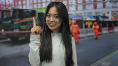 İşçi ve araçlarla dolu kalabalık bir şehir sokağında, gülümseyen ve parmakla işaret eden genç Çinli kadın..