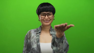 Gözlüklü genç Çinli kadın izole edilmiş yeşil bir arka planda el sıkışma sunuyor, dostluk ve profesyonellik yayıyor, canlı ve modern bir ortamda yakalanıyor..
