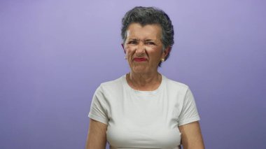 Beyaz tişörtlü yaşlı bir kadın mor arka planda yüzünü buruşturuyor, rahatsızlık ve hoşnutsuzluk anı yakalıyor..