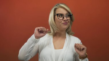 Beyaz bluzlu, gözlüklü, yumruklarını kaldıran ve stüdyoda turuncu arka planda gülümseyen genç sarışın kadın..